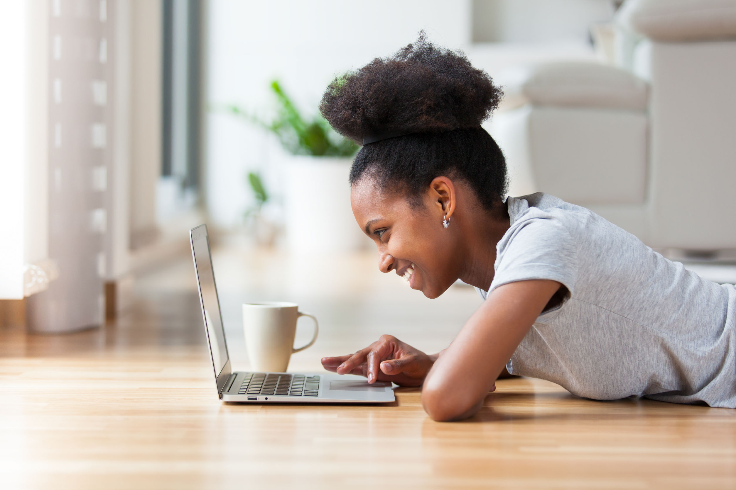 African American girl laying on the floor with her coffee, using her laptop.